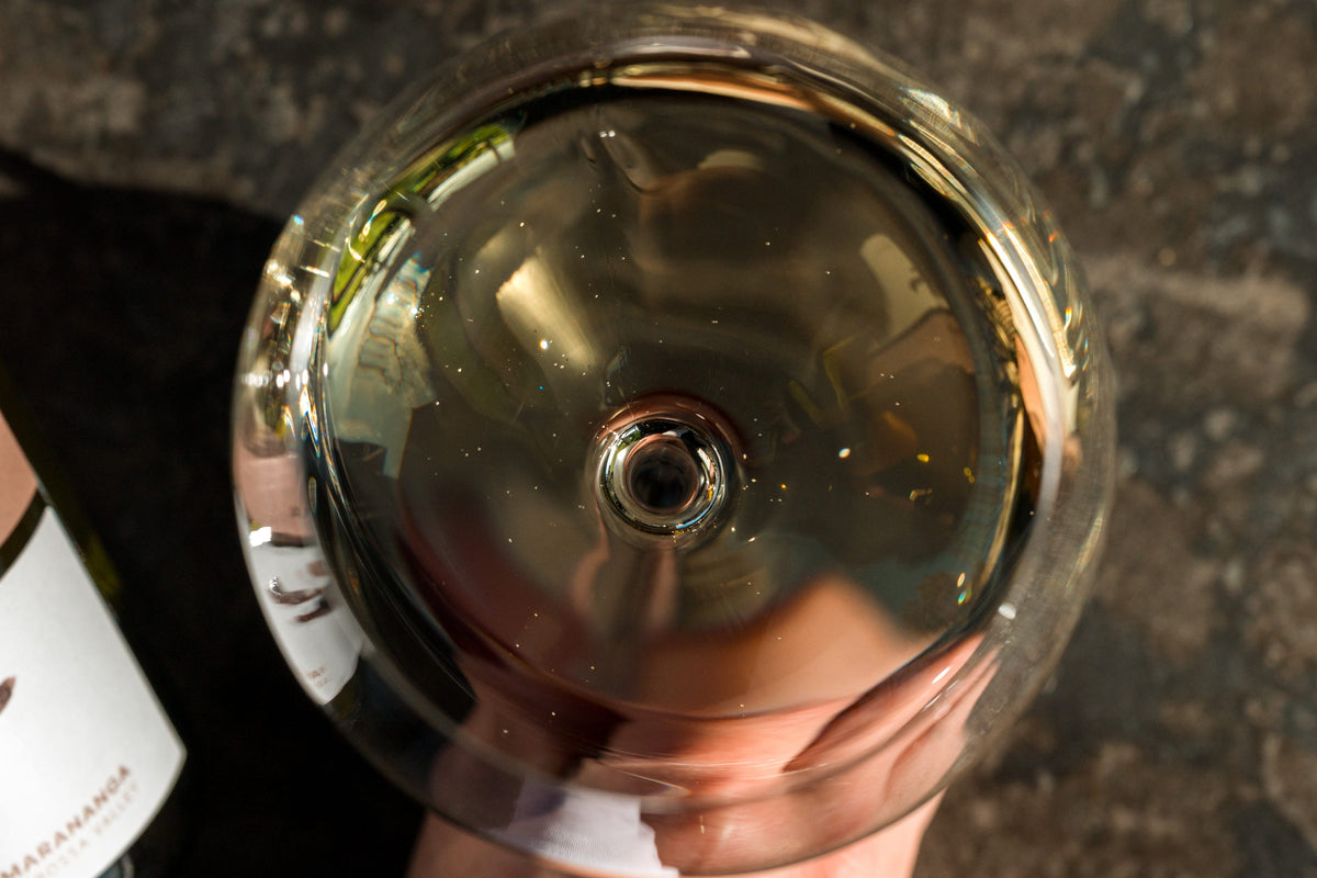 A top-down view of a hand holding a wine glass filled with pale gold wine, with part of a wine bottle visible on the left against a dark textured surface.